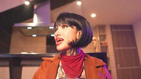 Media: Video of a woman with a bob haircut, wearing a brown jacket and red scarf, smiling in a modern kitchen with stainless steel appliances and purple lighting.