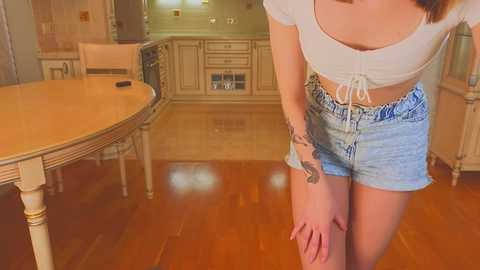 Media: Video of a young woman with light skin, wearing a white crop top and light blue denim shorts, standing in a cozy, wooden-floored kitchen with cabinets and a round table.