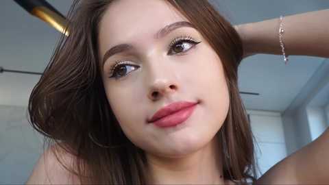 Media: Video of a young woman with fair skin, long brown hair, and well-groomed eyebrows, wearing a delicate silver bracelet. She has a serene expression and is indoors with a modern, minimalist background.