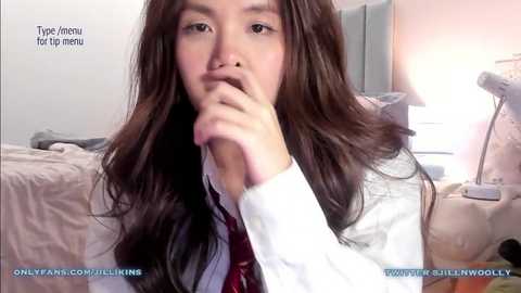 Media: Video of an Asian woman with long, wavy brown hair, wearing a white shirt and maroon tie, sitting on a bed in a softly lit room, thumb to mouth, captioned \"Type Amour for menu.\