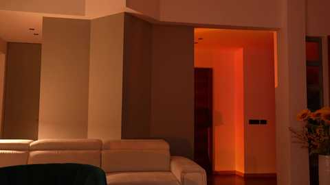 Media: Video of a modern living room with beige walls and a white leather sofa, featuring a warm, orange glow from a nearby doorway, highlighting a stylish, minimalist interior.