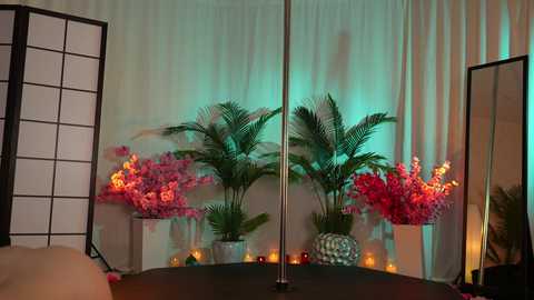 Media: Video of a stage setup with a black pole in the center, surrounded by tall green plants, pink orchids, and warm yellow lighting.