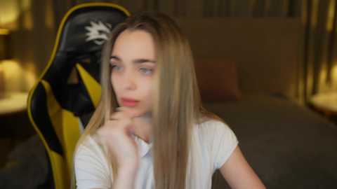 Media: Video of a young Caucasian woman with long, straight blonde hair, wearing a white shirt, sitting on a yellow and black gaming chair, looking introspective.