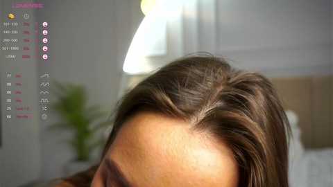 Media: A close-up video of a woman's face with medium brown hair, featuring a detailed view of her scalp and forehead. The background is a blurred bedroom with a bedside lamp and a green plant.