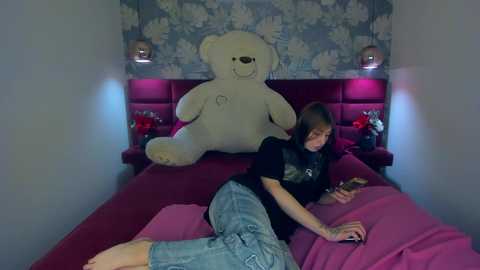 Media: A video of a young woman lying on a pink bed, wearing a black t-shirt and jeans, playing with a smartphone. A large white teddy bear is positioned behind her on the bed, against a floral-patterned wallpaper.