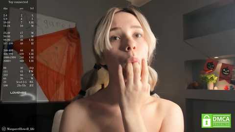 Media: Video of a blonde woman with light skin, wearing a black choker, covering her mouth, standing in a modern room with a calendar and aquarium in the background.