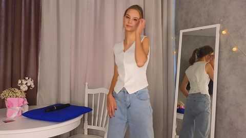 Media: Video of a slim, young woman with light skin, blonde hair, and blue jeans, standing in a room with a white table, a mirror, and gray curtains.