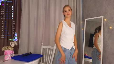 Media: Video of a young woman with light skin and blonde hair, wearing a white sleeveless top and light blue pants, standing in a modern bedroom with a white chair, mirror, and blue cushion.