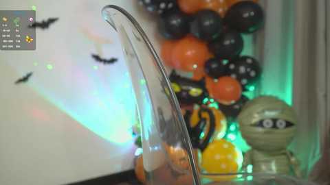 Media: Video of a glass vase filled with orange, black, and yellow balloons; Halloween-themed with black bats on the wall and a green glowing light.