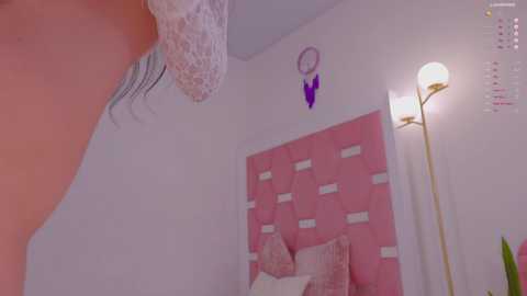 Media: Video of a bedroom with a pink headboard, white pillows, and a dreamcatcher on the wall. A woman in a white lace dress stands near the bed, holding a phone.