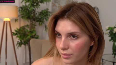 Media: Video of a young woman with fair skin and light brown hair, wearing minimal makeup, sitting in a modern living room with green plants, beige furniture, and a lamp.