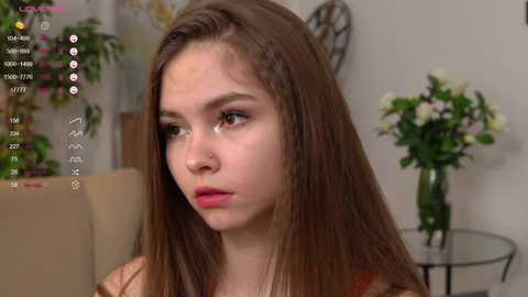 Media: A video of a young, fair-skinned woman with long brown hair, wearing a red top, looking contemplative in a home setting with a vase of flowers.