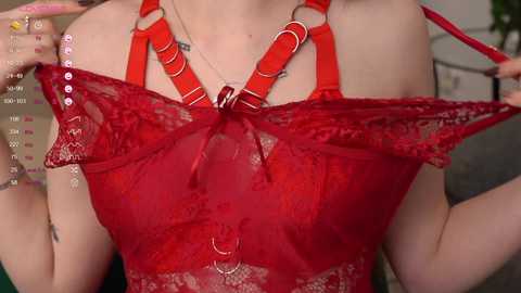 Media: Video of a fair-skinned woman with a slender build wearing a red lace bra with metal rings, revealing her cleavage and a hint of her nipples. Background includes a blurred setting with greenery and a wall.