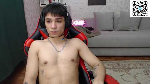 Media: A topless, slender Asian man with short black hair and a silver necklace sits in a red gaming chair. Background shows a grey couch and patterned wallpaper.