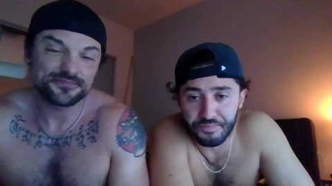 Media: Video of two shirtless men with tattoos, one wearing a black beanie and the other a backward baseball cap, indoors, in a dimly lit room.