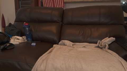 Media: A video of a messy living room couch with a beige blanket, a blue water bottle, and a black hat on the dark brown leather upholstery.