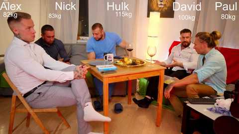 Media: Video of five men seated around a wooden table in a cozy living room, engaging in conversation. Background includes a sofa, windows, and a lamp.