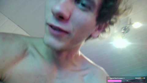 Media: A video of a young, shirtless man with short, curly brown hair and fair skin, taken from a low angle, showing his upper chest and face. The background features a ceiling with recessed lights and a blurred, modern room.