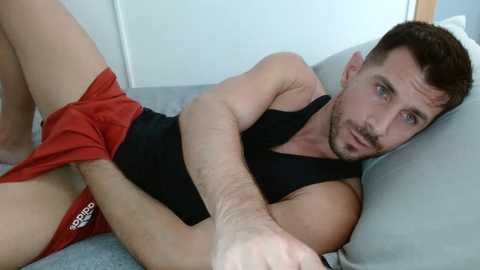 Media: Video of a fit, bearded man in black tank top, red Adidas shorts, lying on a bed with light blue sheets, legs slightly raised.