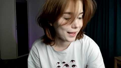 Media: Video of a young woman with shoulder-length, messy brown hair, wearing a white t-shirt with cartoon eyes, smiling softly. Background features a dark curtain and a partially visible doorway.