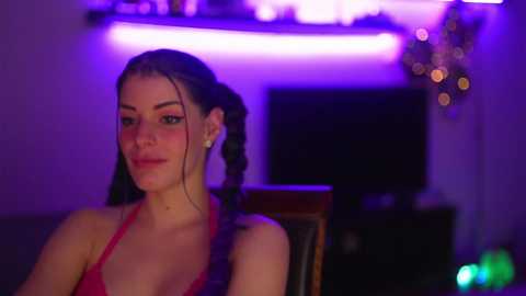 Media: Video of a fair-skinned woman with long, dark braided hair, wearing a red halter top, seated in a dimly lit room with purple lighting. Background includes a TV and Christmas decorations.