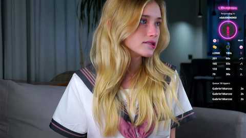 Media: Video of a young blonde woman with wavy hair, wearing a sailor school uniform, sitting indoors; background includes a TV screen displaying a digital interface.