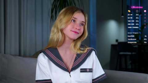 Media: Video of a blonde woman in a white sailor dress, smiling, indoors with dim lighting, potted plants, and a TV displaying weather info in the background.