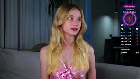 Media: Video of a young Caucasian woman with long, wavy blonde hair, wearing a pink halter top, seated indoors. Background includes a TV screen displaying a fitness app interface.