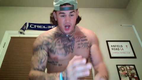 Media: A video of a shirtless man with a tattooed chest, wearing a backwards cap, and a blue bracelet, standing in a room with closed blinds, framed pictures, and a \"Chelseas\" sign.