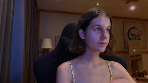 Media: A video of a young woman with shoulder-length dark hair, wearing a floral-patterned spaghetti-strap top, sitting on a black gaming chair in a dimly lit room with beige walls, a lamp, and framed artwork.