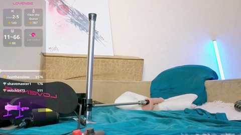 Media: Video of a modern bedroom with a blue bedspread, a standing lamp, and a large abstract painting on the wall. A person is lying in bed, partially obscured by the bedding. The room is cluttered with electronic devices and a laptop.