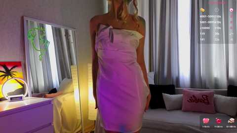 Media: Video of a slender, blonde woman with fair skin, wearing a white towel dress, standing in a dimly lit room with a lamp, palm tree print, and pink pillow.