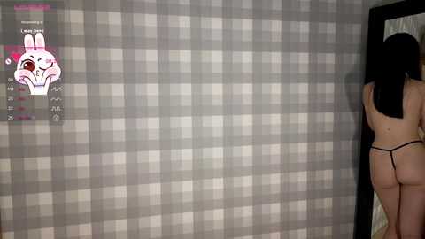 Media: A video of a naked woman with long black hair, wearing only a black thong, standing in front of a gray and white checkered wall, with a virtual rabbit character on the left.