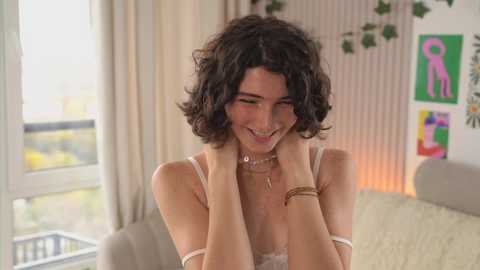 Media: A video of a young woman with curly dark hair, wearing a delicate white dress, grinning broadly while touching her face. The background features a modern, softly lit living room with colorful abstract art.