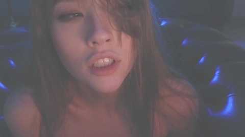 Media: A video of a young woman with long brown hair, slightly tousled, and light skin, wearing a black top. She appears to be in a dimly lit, intimate setting, with a hint of blue light enhancing the moody atmosphere.