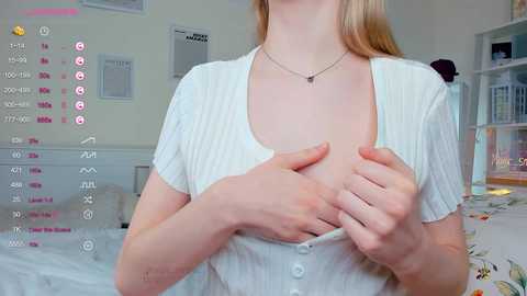 Media: Video of a fair-skinned woman with long blonde hair, wearing a white ribbed top, exposing her breast with her hand. Background shows a neatly arranged room with a bed and a shelf.