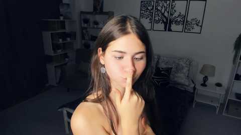 Media: Video of a young woman with long brown hair, light skin, and a slender physique, sitting in a dimly lit bedroom. She has a thoughtful expression, touching her finger to her lips. The room features a bed with a white lamp and framed tree art on the wall.