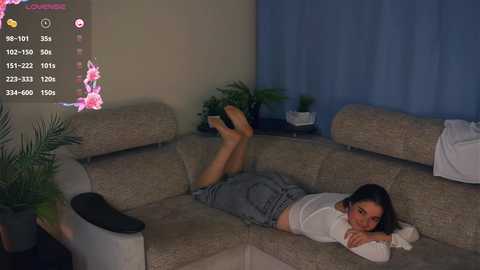 Media: A video of a young woman lying on a beige sectional sofa, wearing a white shirt and grey shorts, with her legs kicked up, in a dimly lit living room.