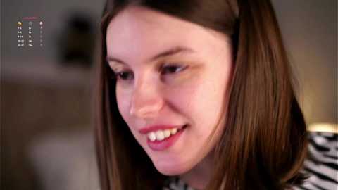 Media: Video of a smiling Caucasian woman with straight, shoulder-length brown hair, wearing a zebra-print top, in a softly lit indoor setting.