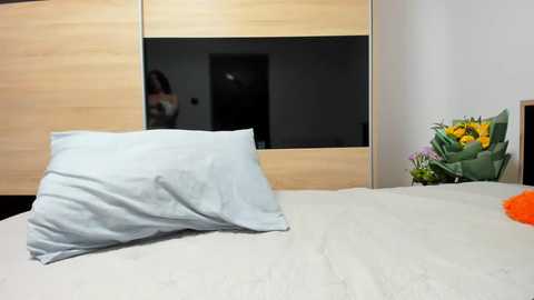 Media: A video of a neatly made bed with a white pillow and light blue duvet, featuring a wooden headboard with a black glass panel. To the right, a vase of vibrant yellow flowers sits on a white bedside table.