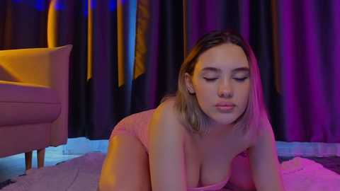 Media: A video of a young Caucasian woman with light skin, straight blonde hair, and wearing a pink dress, lying on the floor with her eyes closed, in a dimly lit room with purple and yellow lighting.