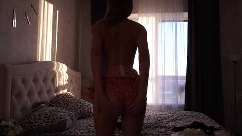 Media: Video of a woman standing with her back to the camera in a dimly lit bedroom, wearing red and white polka dot panties, sunlight casting shadows on her bare back.