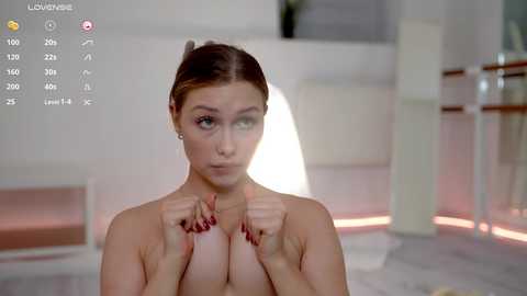 Media: Video of a nude, light-skinned woman with medium-sized breasts, brown hair in a bun, hands covering her chest, standing in a minimalist room with white walls and furniture.