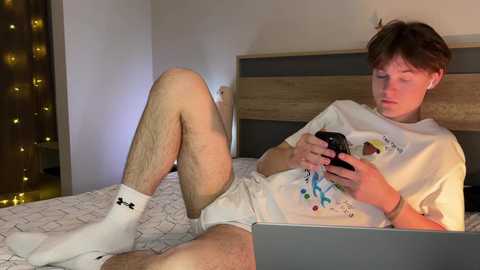 Media: Video of a young, fair-skinned, slender, Caucasian man with short brown hair, wearing white Under Armour socks and a white t-shirt, playing a video game on a laptop, lying on a bed with a wooden headboard in a dimly lit room.