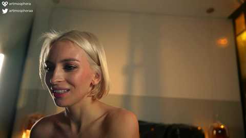 Media: Video of a smiling, topless blonde woman with fair skin and short hair, in a dimly lit room with warm lighting, blurred background, and a watermark.