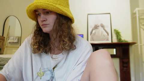 Media: Video of a young woman with curly brown hair, wearing a yellow knit beanie and a white T-shirt, sitting in a bedroom with a wooden dresser, framed photo, and a mirror in the background.