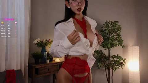 Media: A video of a curvy Asian woman with long black hair, red lipstick, and glasses, wearing a white unbuttoned shirt and red lace lingerie, standing in a dimly lit room with a tall lamp, wooden desk, and potted plants.