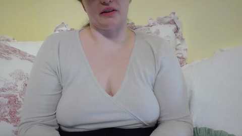 Media: Video of a middle-aged woman with fair skin, wearing a light grey V-neck top, revealing ample cleavage, seated on a bed with floral-patterned pillows and a yellow wall in the background.