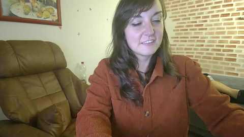 Media: Video of a smiling woman with long brown hair, wearing an orange sweater, seated on a brown leather recliner in a cozy living room with brick wall and framed artwork.