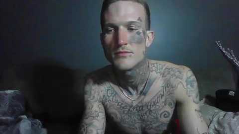 Media: Video of a shirtless, pale-skinned man with short brown hair and numerous intricate tattoos covering his chest and arms, gazing thoughtfully at the camera against a dark, blurry background.